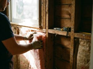 Werker installeert roze glaswol isolatie tussen houten balken in huismuur met gereedschap op bouwplaats