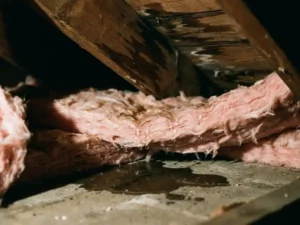 Doorsnede dakisolatie met waterdruppels op roze glaswol, vochtvlekken op houten balken en plas water op betonvloer
