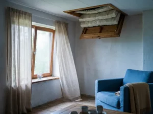 Zonnige Nederlandse woonkamer met dikke dakisolatie zichtbaar via luik, linnen gordijnen bij gekanteld raam en glas koud water op houten vensterbank.